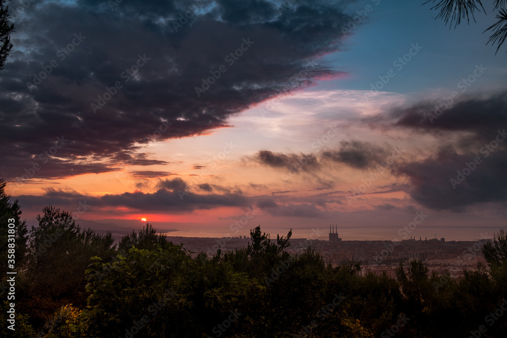 dramatic sunrise view of barcelona at dusk from guinardo park, with sun rising through clouds and cityscape with tres xemeneies