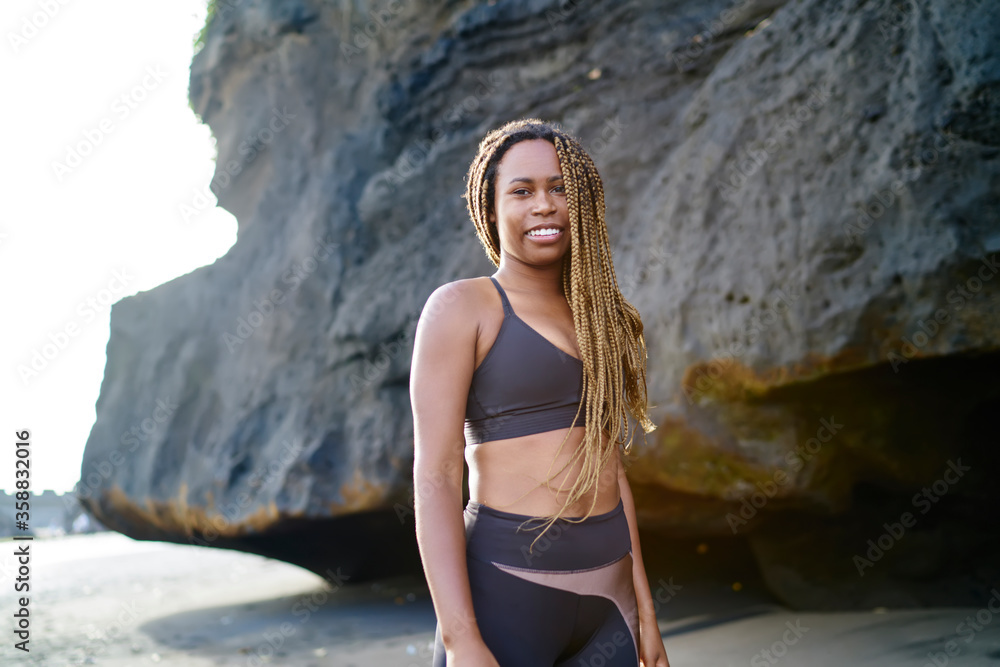 Portrait of cheerful dark skinned sportwoman dressed in active clothes smiling at camera.Positive afro american young woman in tracksuit standing outdoors on coastline before morning workout Portrait of cheerful dark skinned sportwoman dressed in active clothes smiling at camera.Positive afro american young woman in tracksuit standing outdoors on coastline before morning workout