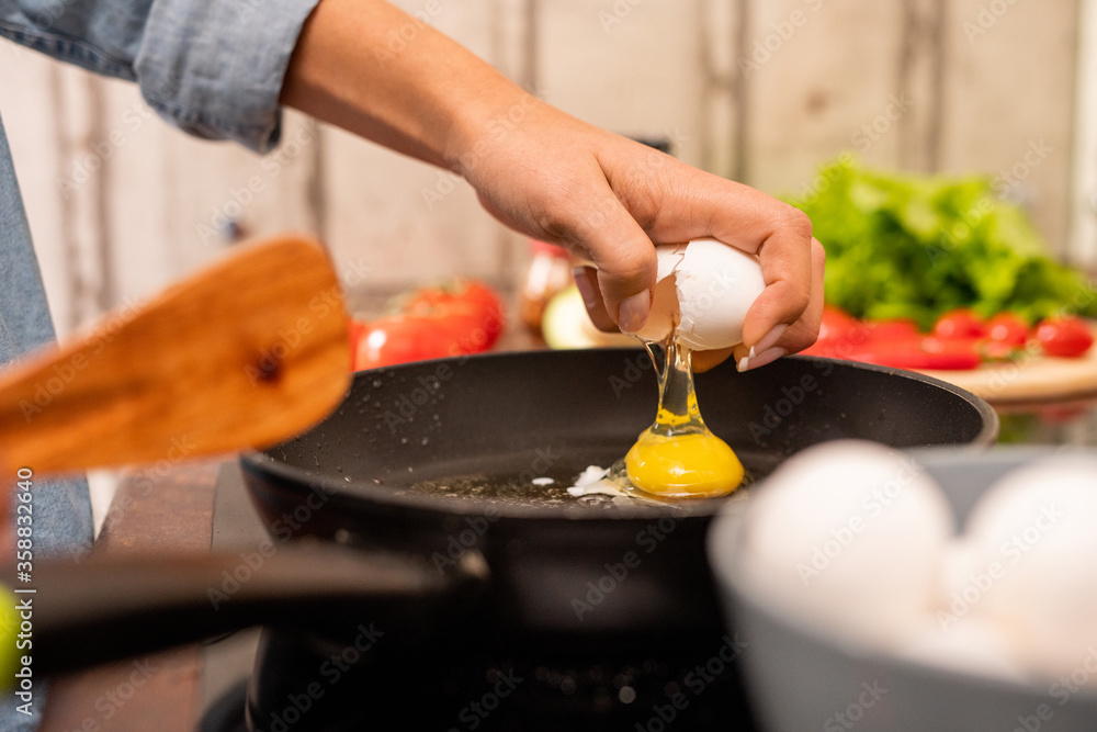 Hand of young woman breaking fresh egg on frying pan standing on electric stove