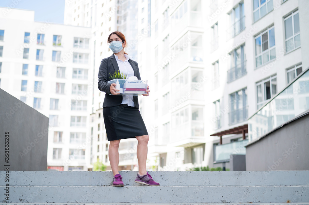 Depressed woman wearing a medical mask walking down the street with a box of personal stuff. Female office worker in a suit and sneakers. Economic crisis and unemployment during the epidemic covid 19.
