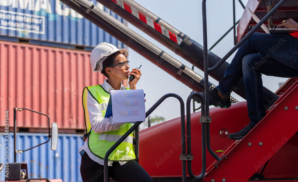 Woman Foreman in hard hat and safety vest holding clipboard checklist and talks on two-way radio, Engineer team control loading containers box from cargo