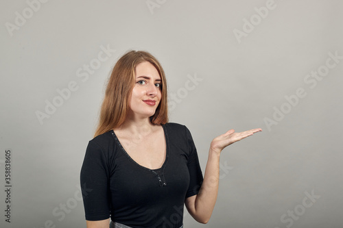 Palms up, place your product here, raising arms wide spread is catching something. Young attractive woman, dressed black t-shirt, grey background