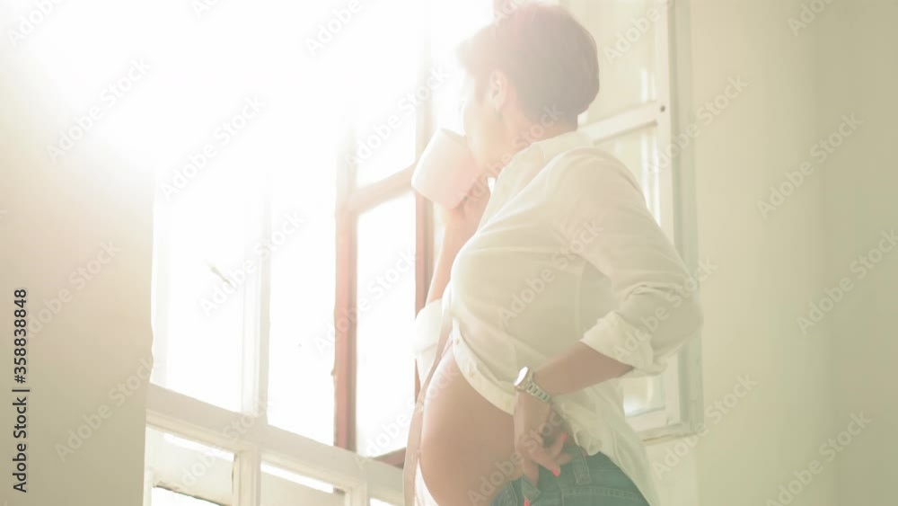 Young woman come closer to a window expecting a baby drinking tea. Young pregnant woman resting touching her belly standing at the window. Happy pregnant caucasian woman at home. Prores 422. Young woman come closer to a window expecting a baby drinking tea. Young pregnant woman resting touching her belly standing at the window. Happy pregnant caucasian woman at home. Prores 422.