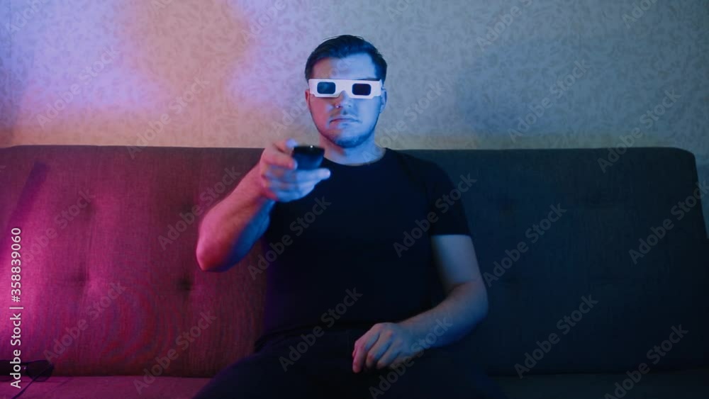 A man in stereo glasses sits on the couch and switches channels on the TV. A man in stereo glasses sits on the couch and switches channels on the TV.