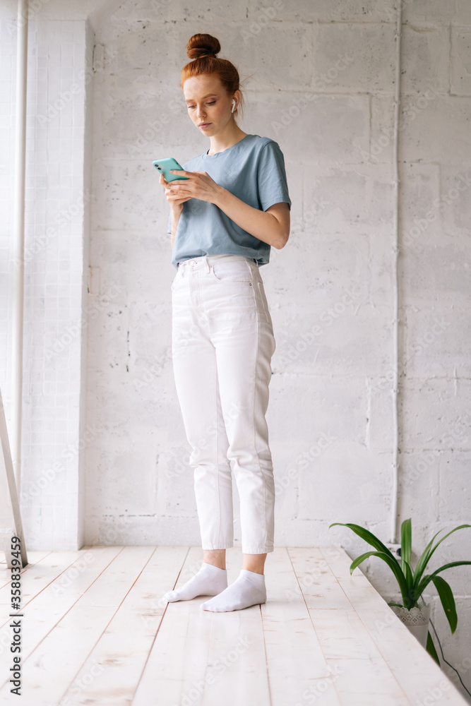 Beautiful redhead young woman is using cell phone while standing near big panoramic window. Pretty girl is typing message on cellphone on white background. Beautiful redhead young woman is using cell phone while standing near big panoramic window. Pretty girl is typing message on cellphone on white background.