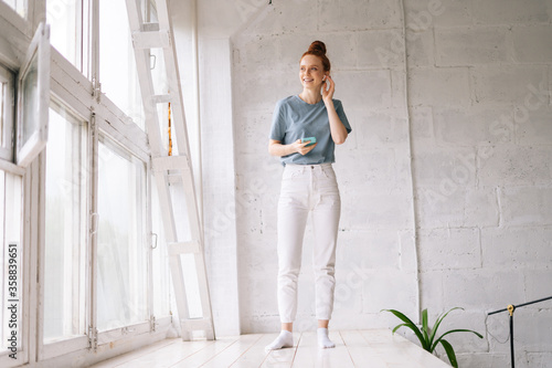 Cheerful redhead young woman is using cell phone while standing near big panoramic window. Cute lady wearing wireless earphones is listening relaxed pleasant music at home office on white background.