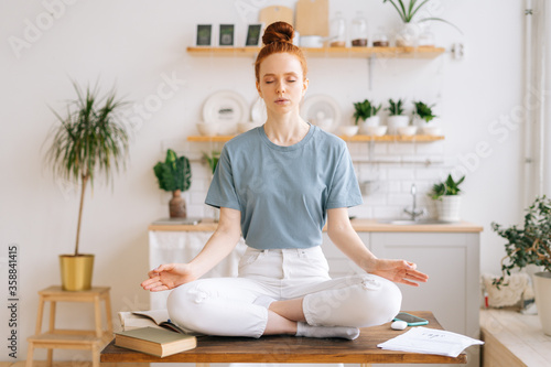 Peaceful redhead young woman is sitting with closed eyes on table in lotus position and makes deep breath-exhalation at home office. Peaceful female sitting in lotus pose.