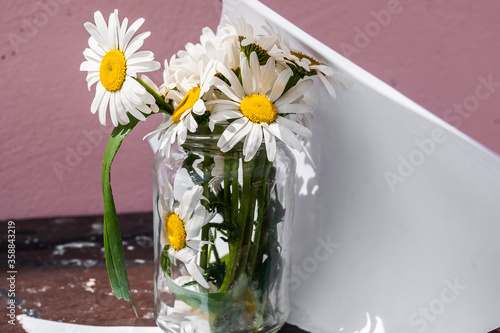 Fototapeta Naklejka Na Ścianę i Meble -  Torn pieces of white paper and a bouquet of daisies in a glass jar on a wooden background against a pink wall. The concept of cleanliness, skin care, cosmetics and perfume.