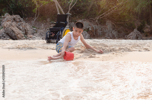 Asian special child playing sand and crawls happily on the beach near the wheelchair,Blue sea background,Natural therapy,Life in the education age of disabled children,Happy disability kid concept.