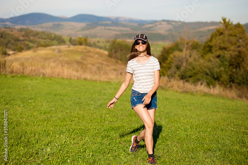 Carefree happy woman in sunglasses, cap and jeans shorts walk on green grass meadow on top of mountain enjoying nature. Freedom