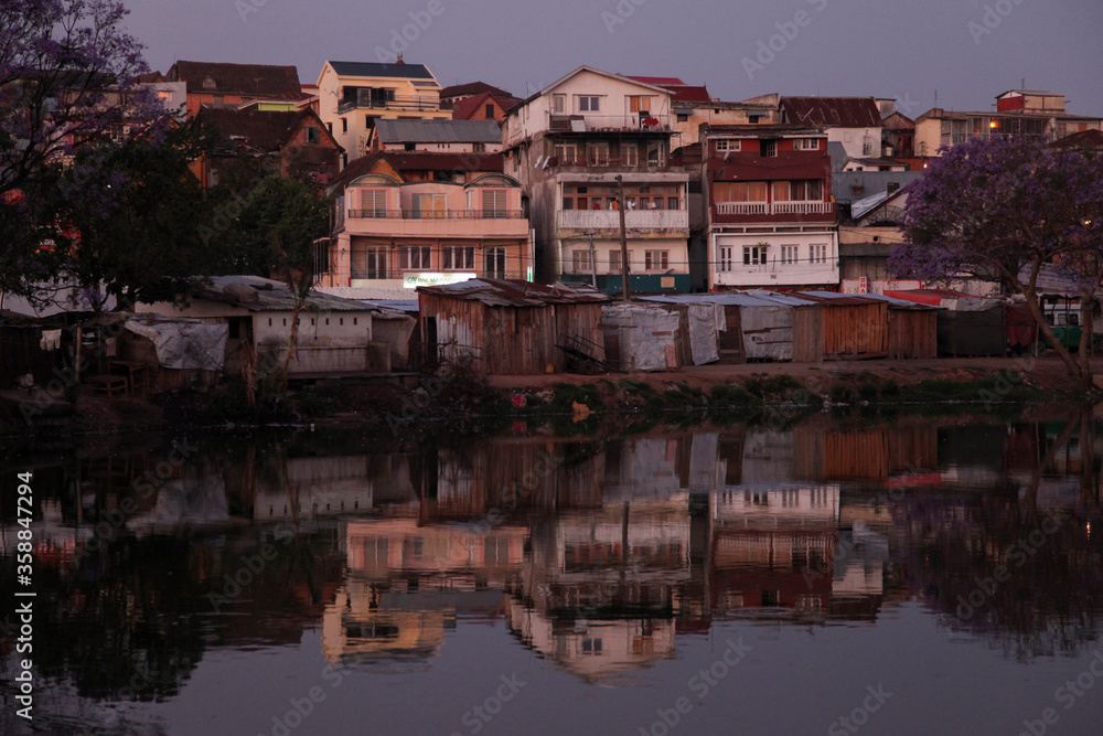 Obraz premium Houses and buildings reflect in the still waters of a lake in Antananarivo, Madagascar just after sunset.