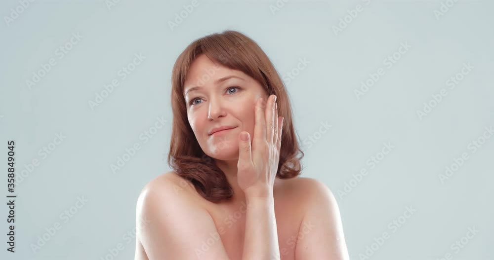 Pretty mature female applies skin care product by touching face with hand, close up, spa and wellness concept. Woman applying anti-age cream on her skin isolated on white background. Prores 422. Pretty mature female applies skin care product by touching face with hand, close up, spa and wellness concept. Woman applying anti-age cream on her skin isolated on white background. Prores 422.