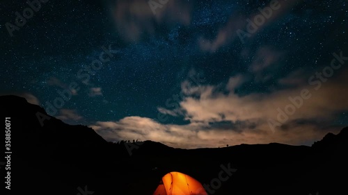 Orange tent with a burning light inside stands in a clearing under the starry sky. On the horizon mountains and forest. Concept of a beautiful starry night sky and the Milky Way. Night in the mountain