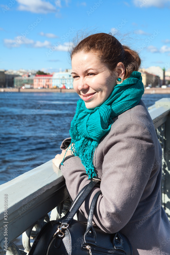Fototapeta premium Smiling adult woman wearing coat standing on bridge, looking at the distance, sunny weather is in a city