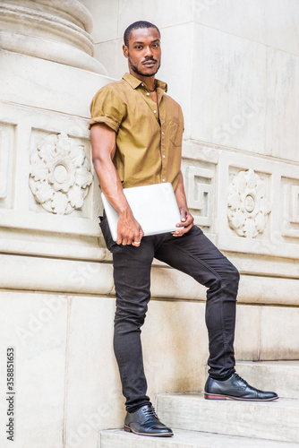 Young African American Male College Student with beard studying in New York, wearing green short sleeve shirt, black pants, leather shoes, carrying laptop computer, standing against white marble wall.