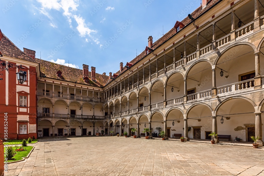 Opocno / Czech Republic - June 16 2020: View of the castle courtyard with arcades and red facade. Stone paving in the foreground. Sunny summer day with blue sky and white clouds.