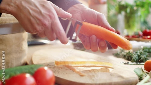 Close-up view of hands of male cook peeling fresh carrot with vegetable peeler while preparing food at kitchen table photo