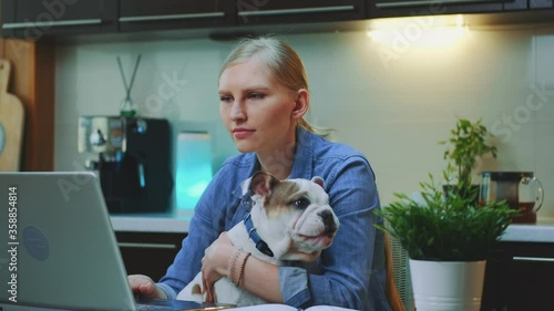 Close-up shot of cheerful woman hugging small dog in the kitchen. She working on the computer and holding small dog on her hands