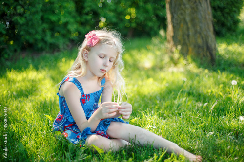 Pensive young girl in blue dress sitting on ground outdoors on summer sunny day. Cute pretty little child kid dreaming thinking on meadow outside.  Authentic candid childhood lifestyle. © anoushkatoronto