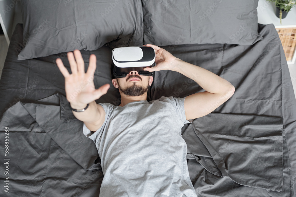 Above view of young bearded man immersed into virtual reality lying on bed and watching video in VR goggles Above view of young bearded man immersed into virtual reality lying on bed and watching video in VR goggles