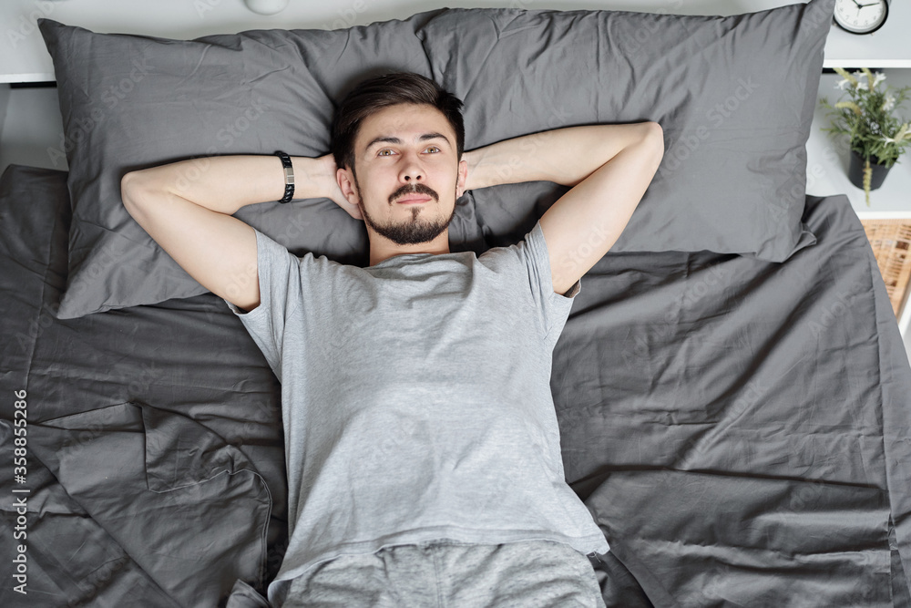 Above view of pensive young bearded man holding hands behind head while relaxing in bed during quarantine