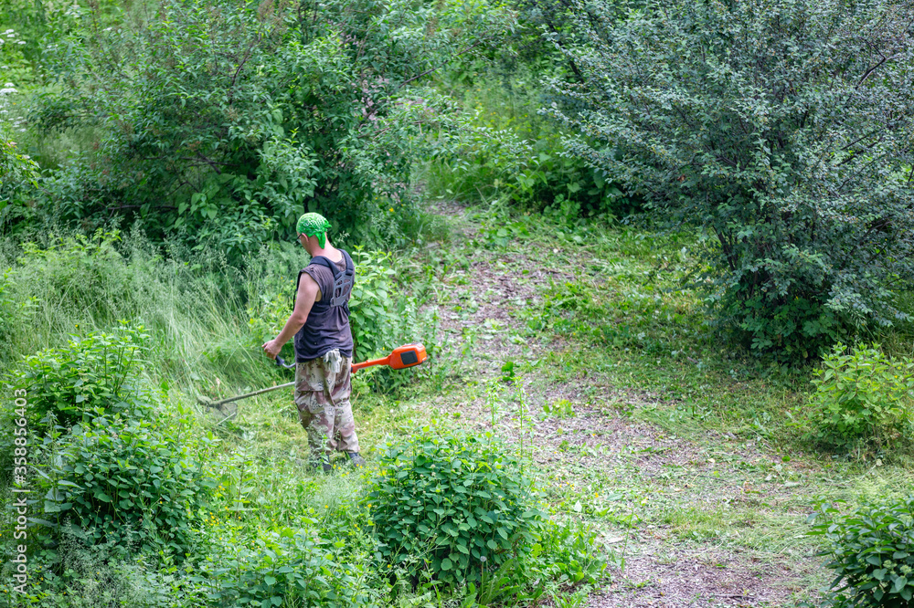 An adult man in uniform with an electric lawn trimmer cuts a lawn in the city park on a summer day An adult man in uniform with an electric lawn trimmer cuts a lawn in the city park on a summer day