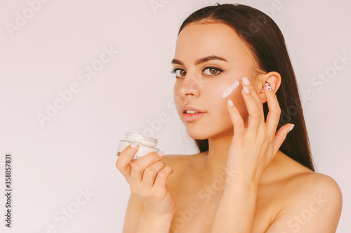 Portrait beautiful young woman apply moisturizer face lotion isolated white background. Attractive sensual girl use face cream from jar for healthy smooth skin. Beauty treatment, body care cosmetics