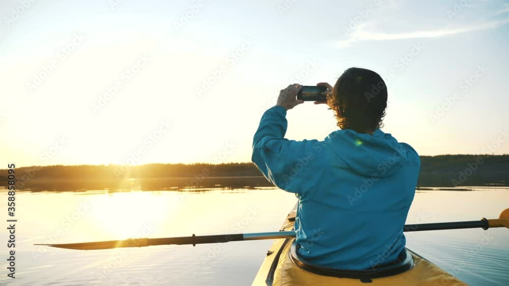 man makes a sunset photo on a smartphone sitting in a kayak on a calm lake, outdoor activities man makes a sunset photo on a smartphone sitting in a kayak on a calm lake, outdoor activities