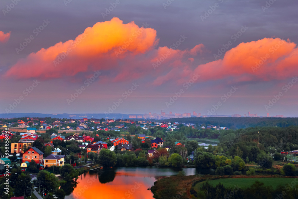 Beautiful dramatic landscape, bright pink clouds reflected in river, country houses in light of dawning sun and distant city in Sloboda village, Moscow region, Russia