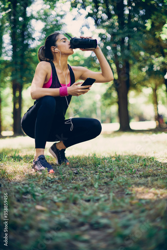 Young attractive caucasian sportswoman crouching, holding smart phone and drinking refreshment. Running in nature concept.