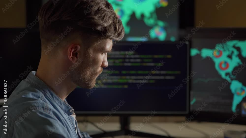 Side view of young man thinking with his hand on chin surrounded by multiple computer monitors with stock market data sitting at desk in dark office Side view of young man thinking with his hand on chin surrounded by multiple computer monitors with stock market data sitting at desk in dark office