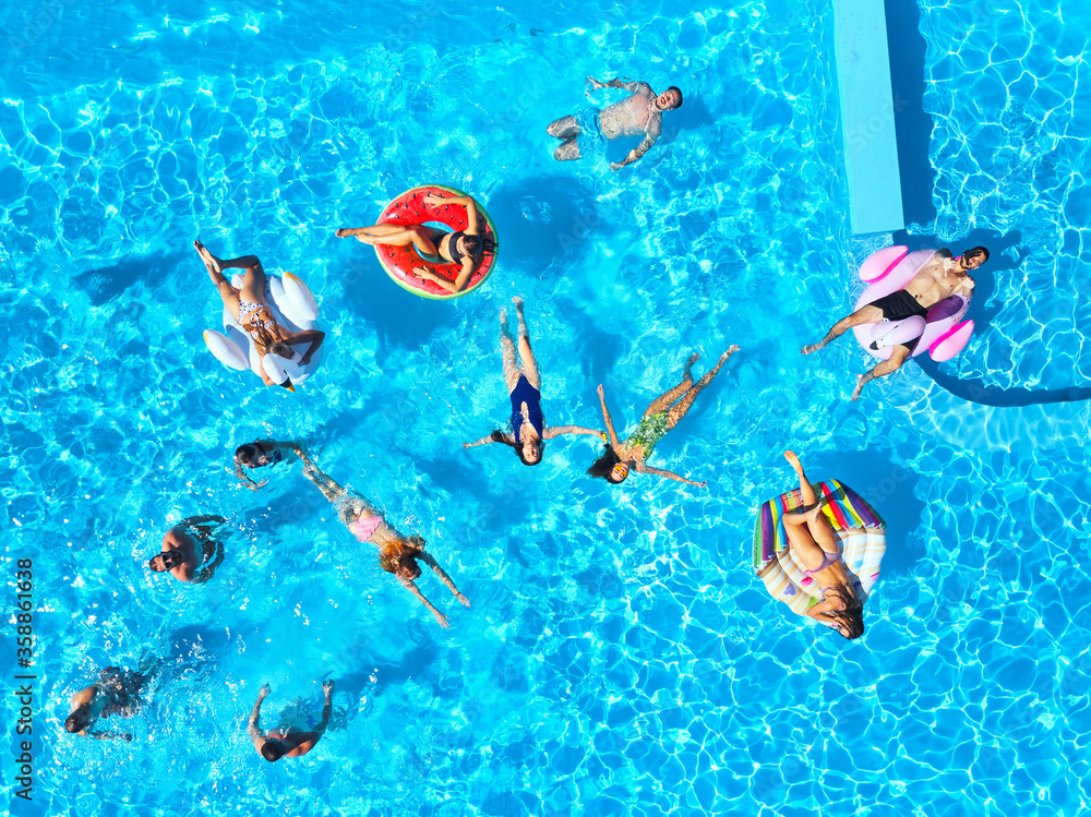 Aerial of friends having party in swimming pool with inflatable flamingo, swan, mattress. Happy young people relax at luxury resort on sunny day. View from above. Girls in bikini sunbathing in a sun.