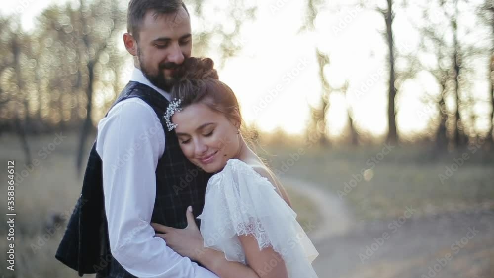 Stylish young bride and groom hug and enjoy solitude in nature. Happy european wedding couple resting outside on a romantic evening against the background of blurry sun and trees. Active rest outside. Stylish young bride and groom hug and enjoy solitude in nature. Happy european wedding couple resting outside on a romantic evening against the background of blurry sun and trees. Active rest outside.