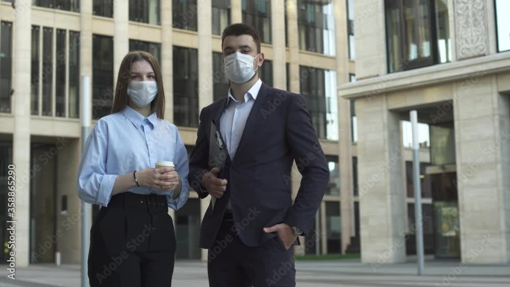 Young businessman in dark blue suit and businesswoman in formal suit are wearing medical masks and looking in the camera near modern office building during pandemic covid-19 coronavirus quarantine.
