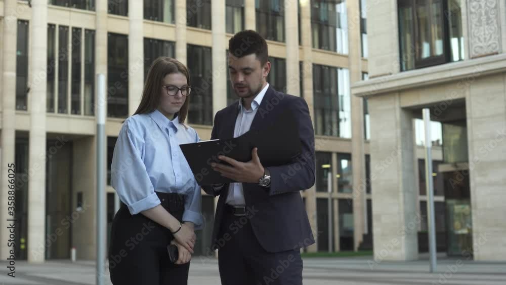 Young businessman in dark blue suit show his businesswoman colleaguein formal suit documents and talk about work near modern office building. Young businessman in dark blue suit show his businesswoman colleaguein formal suit documents and talk about work near modern office building.