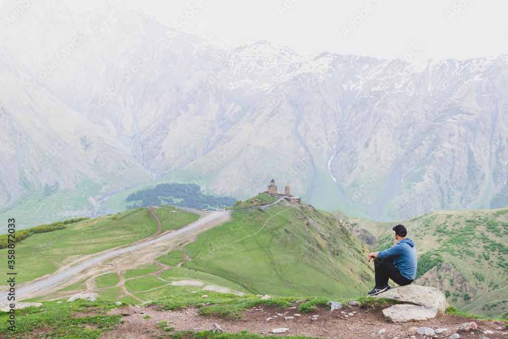 Male tourist in casual clothes with sits relaxed and looks to Gergeti trinity church surounded by green nature. Solo travel in Kazbegi national park.