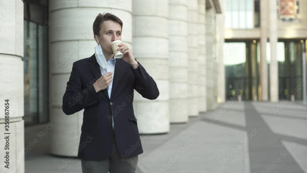 Young businessman in dark blue suit takes off his medical mask near modern office building during pandemic covid-19 coronavirus to take a sip of coffee and then puts it back on. Young businessman in dark blue suit takes off his medical mask near modern office building during pandemic covid-19 coronavirus to take a sip of coffee and then puts it back on.