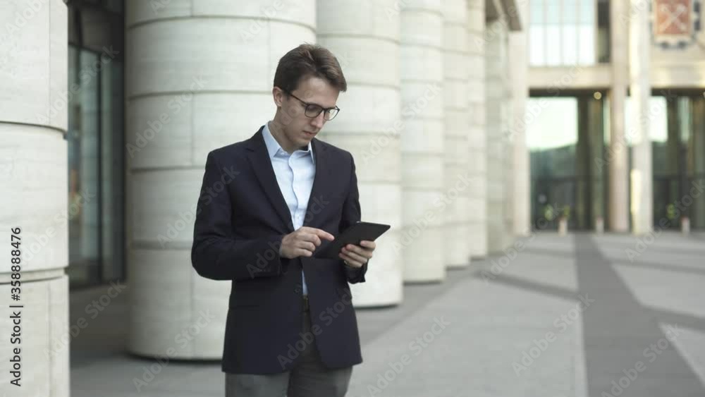 Young businessman in dark blue suit and glasses checking his tablet on the street near modern office building and look away. Young businessman in dark blue suit and glasses checking his tablet on the street near modern office building and look away.