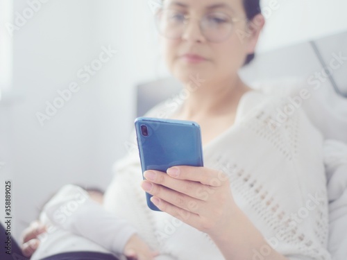 Madre revisa el celular mientras da de comer a su bebé en una cama. Mamá escribe en el celular mientras amamanta a su hijo recién nacido.