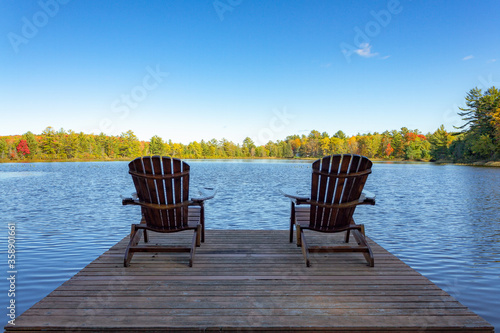 Fototapeta Naklejka Na Ścianę i Meble -  Two Muskoka chairs sitting on a wood dock facing a lake in a calm autumn season sunny day, with the chairs in the sun shadow.