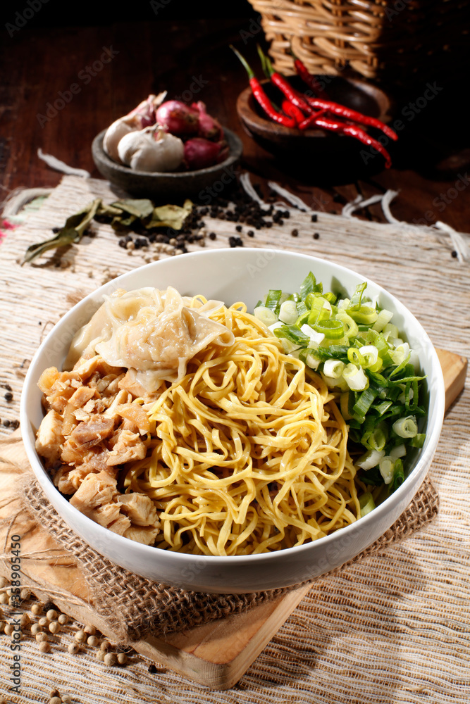 Yamin noodles with boiled chicken meat and boiled dumplings and leeks, Isolated on a white bowl with a burlap sack placemats and cutting board, with a background of cooking spices.