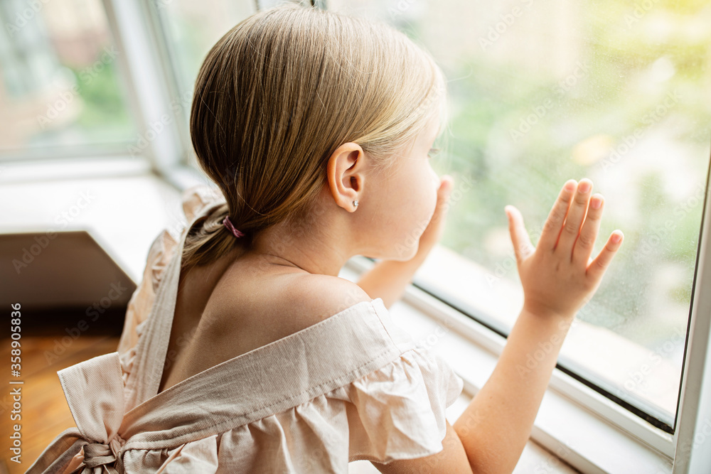 Φωτογραφία Stylish little girl with blonde hair sitting at home near window during coronavirus covid-19 self isolation