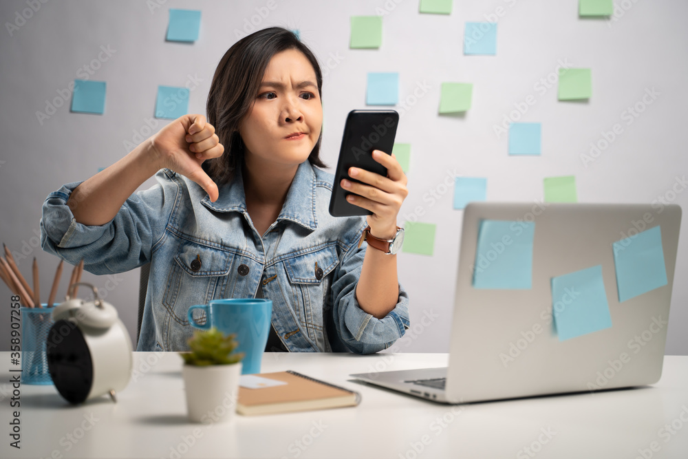 Asian woman boring looking at smart phone reading news showing thumb down at home office. WFH. Work from home. Prevention Coronavirus COVID-19 concept. Asian woman boring looking at smart phone reading news showing thumb down at home office. WFH. Work from home. Prevention Coronavirus COVID-19 concept.