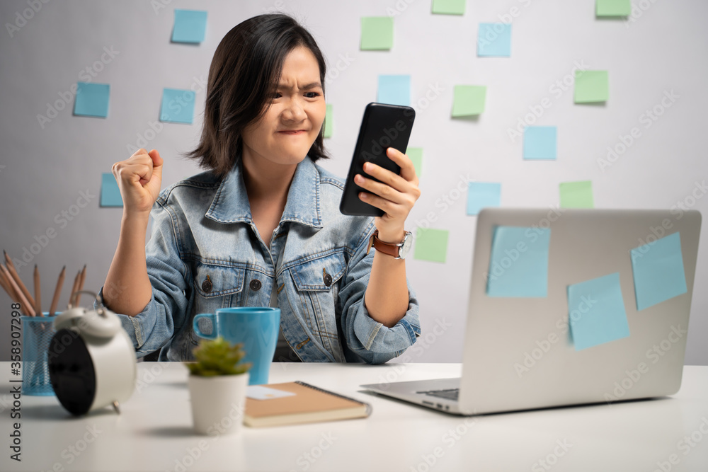 Asian woman angry looking at smart phone reading news at home office. WFH. Work from home. Prevention Coronavirus COVID-19 concept. Asian woman angry looking at smart phone reading news at home office. WFH. Work from home. Prevention Coronavirus COVID-19 concept.