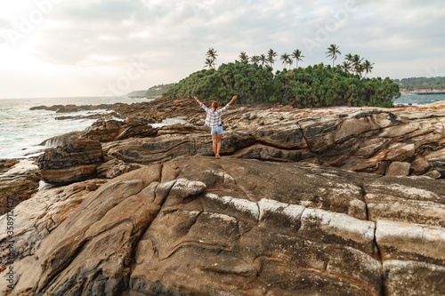 Successful woman outstretched arms on seaside rock cliff edge. Woman with raised arms stand on the rocks with tropical landscape