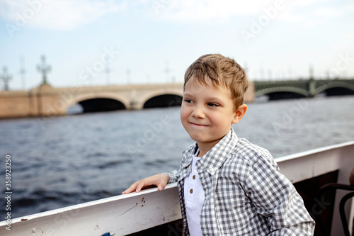 cute caucasian boy looking aside wearing henley tee and shirt on a river boat tour along rivers and channels of Saint Petersburg. Trinity Bridge on background. Image with selective focus 