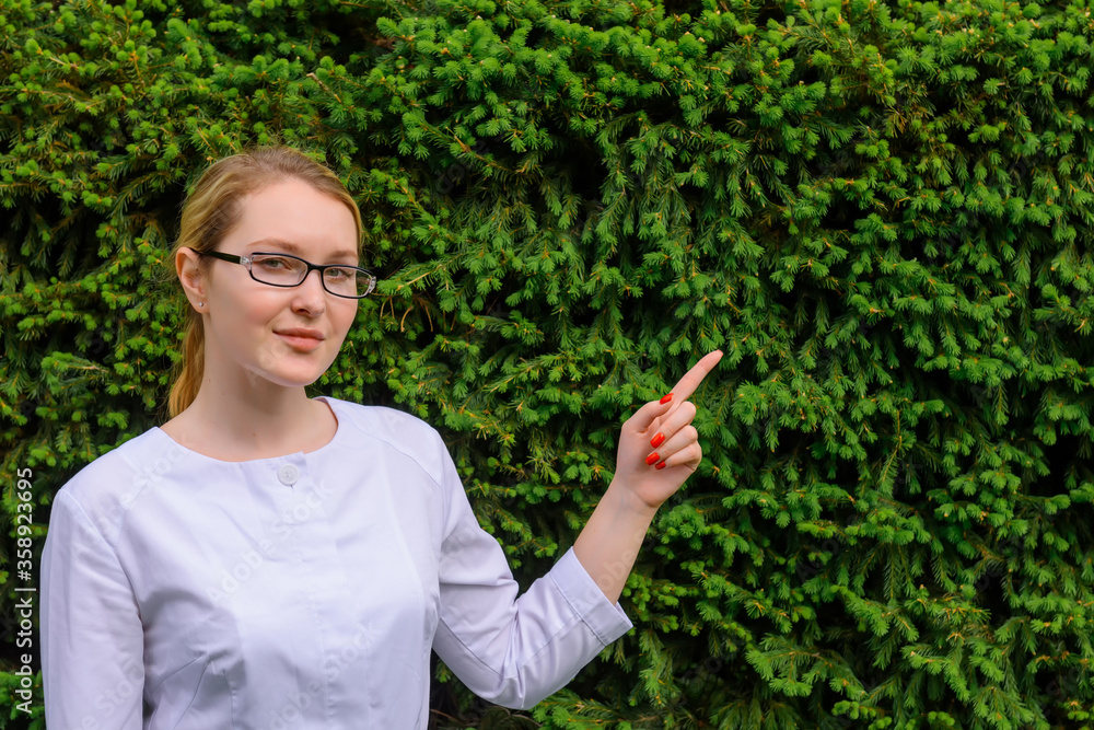 Female doctor with finger point up. Nutritionist in white coat and glasses on green foliage background with copy space. Image for advertising scientific developments in the food and medical industry. Female doctor with finger point up. Nutritionist in white coat and glasses on green foliage background with copy space. Image for advertising scientific developments in the food and medical industry.