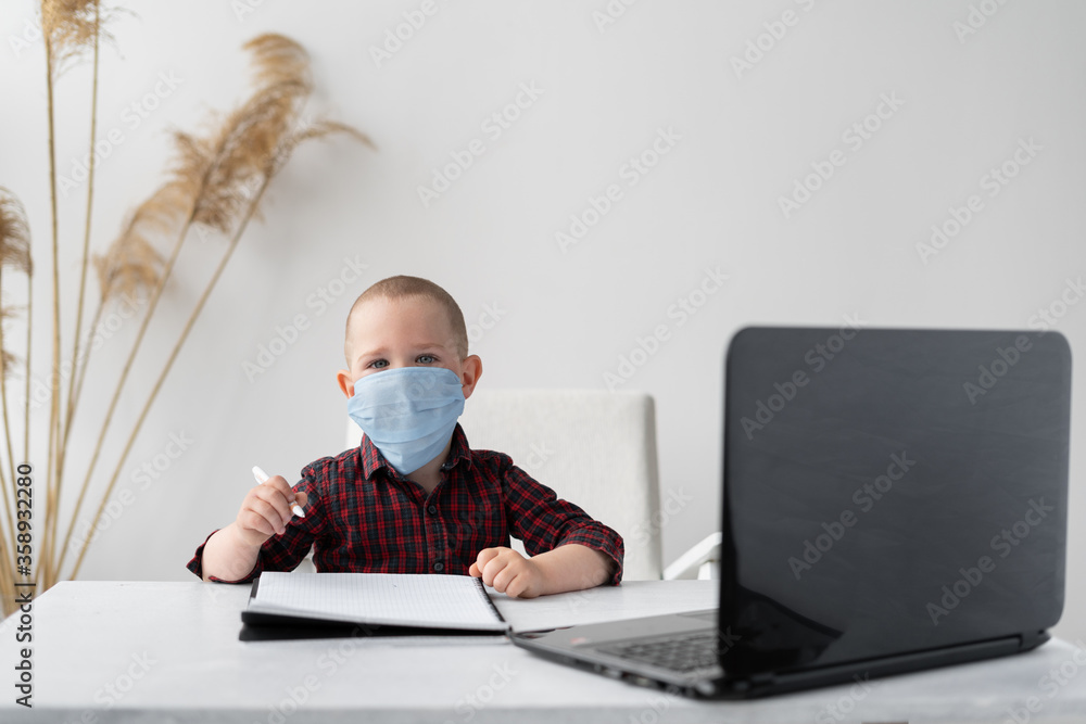 Home schooling education. Online education. A boy sits and does homework at a laptop. He has a pen in his hands and there is a notebook on the table. A medical mask is on the face. Home schooling education. Online education. A boy sits and does homework at a laptop. He has a pen in his hands and there is a notebook on the table. A medical mask is on the face.
