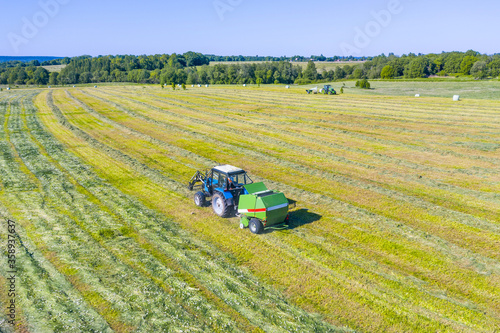 An agricultural tractor collects mowed grass for agricultural use and wraps hay bales in a plastic field, aerial view.