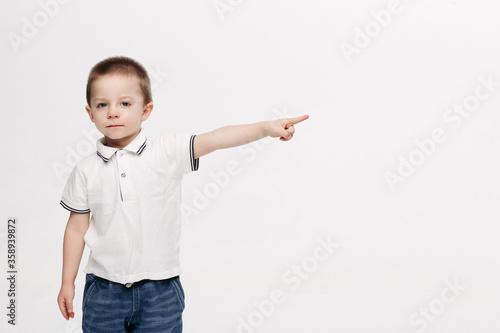 Little cute boy wearing in white polo t-shirt, jeans posing at studio pointing by finger up and looking at stars. Stylish kid dreaming and rising up. Child mood. Kids fashion. White studio background.